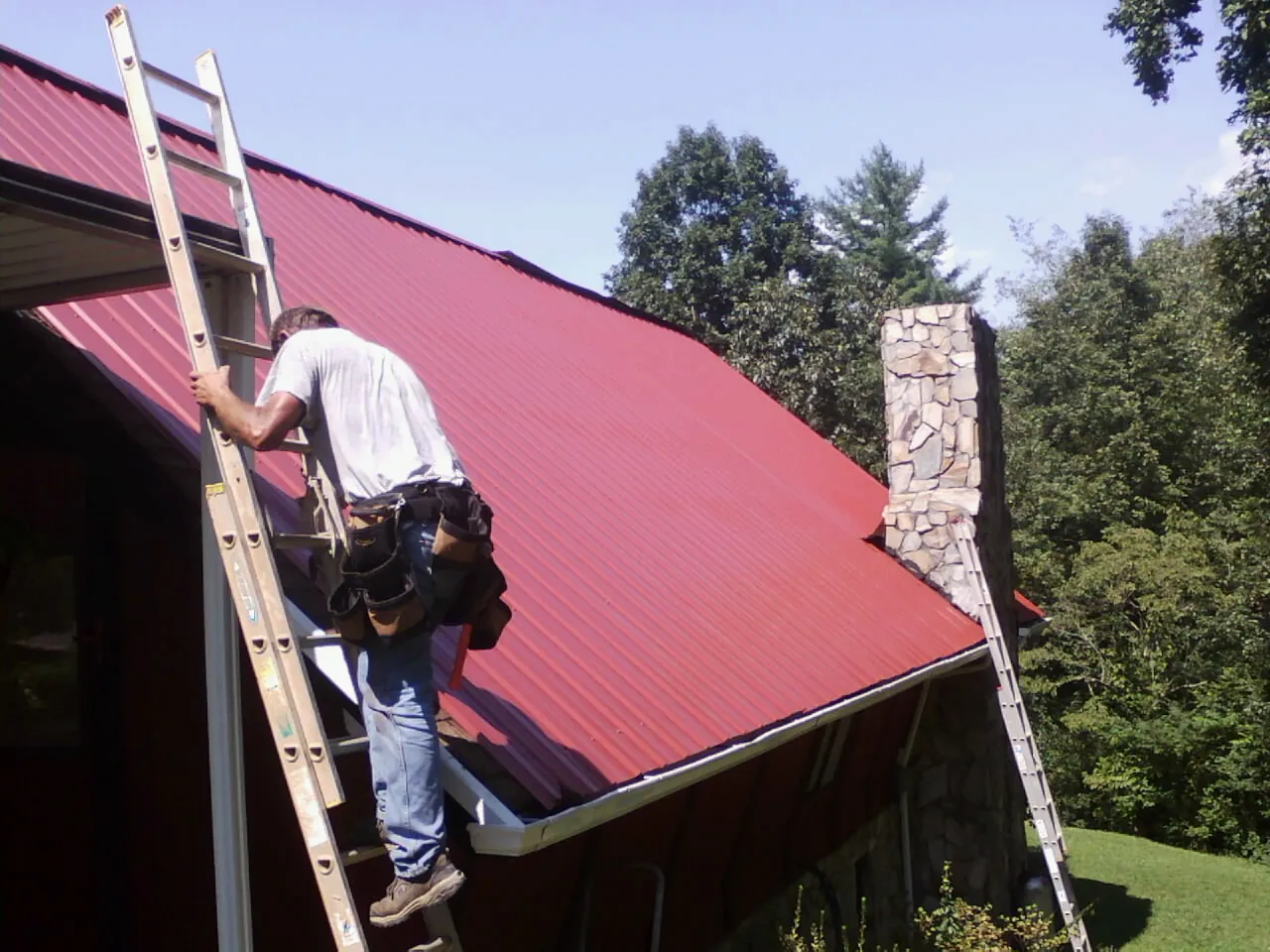 Roofer on a ladder beside a red metal roof and stone chimney — Soffit & Fascia Repair by Roofing Brothers in New Bloomfield